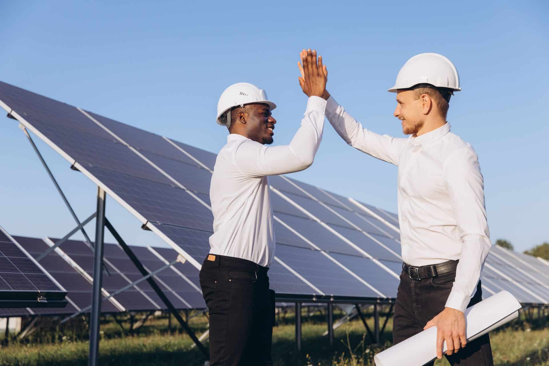 Two engineers in hard hats high-fiving near solar panels in a garden.