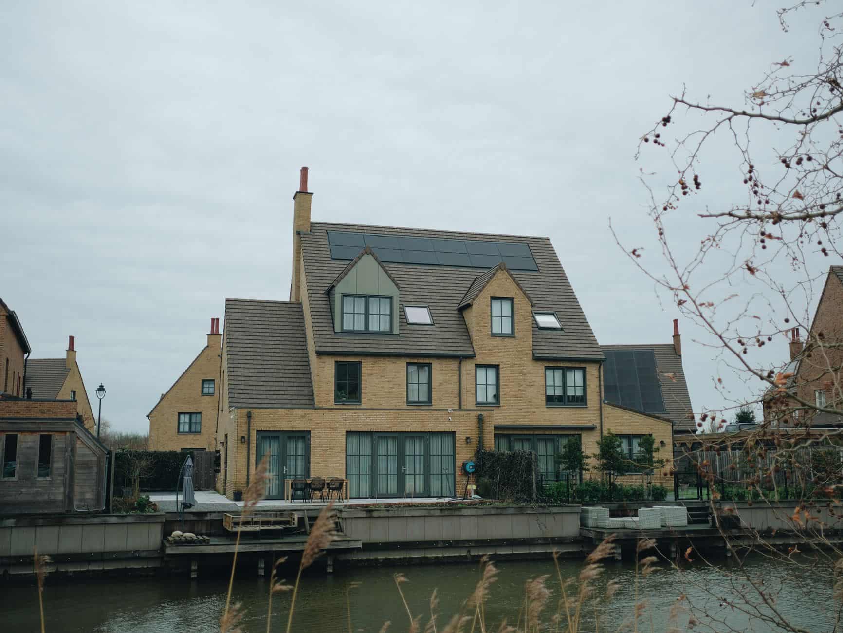 Modern house with solar panels on roof near water under cloudy sky.