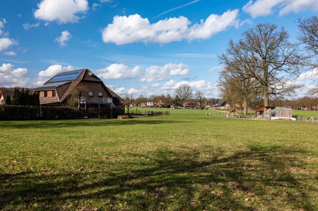 Spacious green field with a house featuring solar panels under a blue sky.