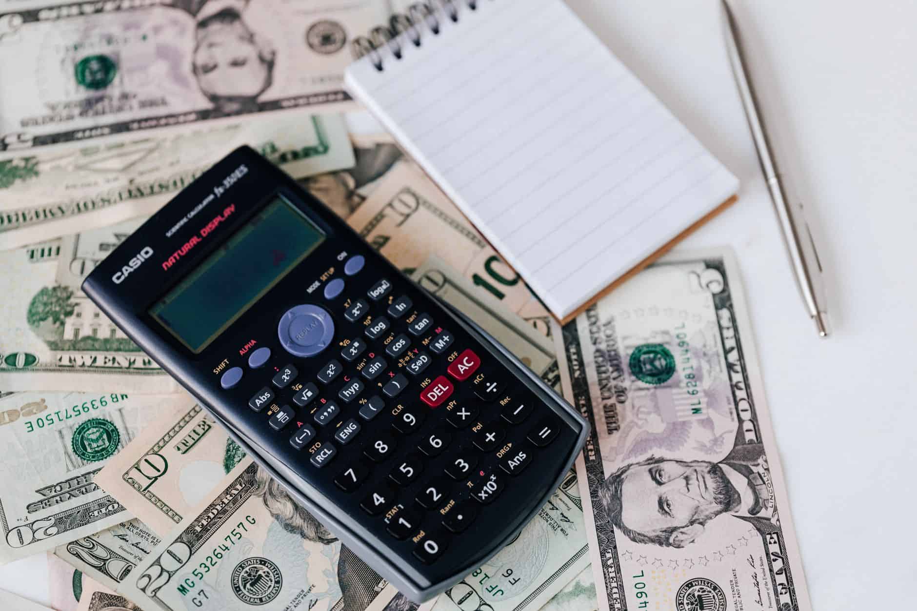 From above electronic calculator and notepad placed over United States dollar bills together with metallic pen for budge