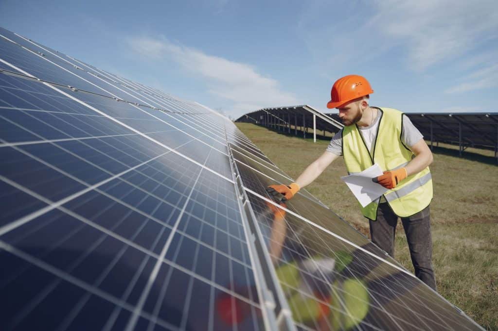 Young bearded male engineer in protective helmet and heat resistant gloves touching solar panel while standing on field