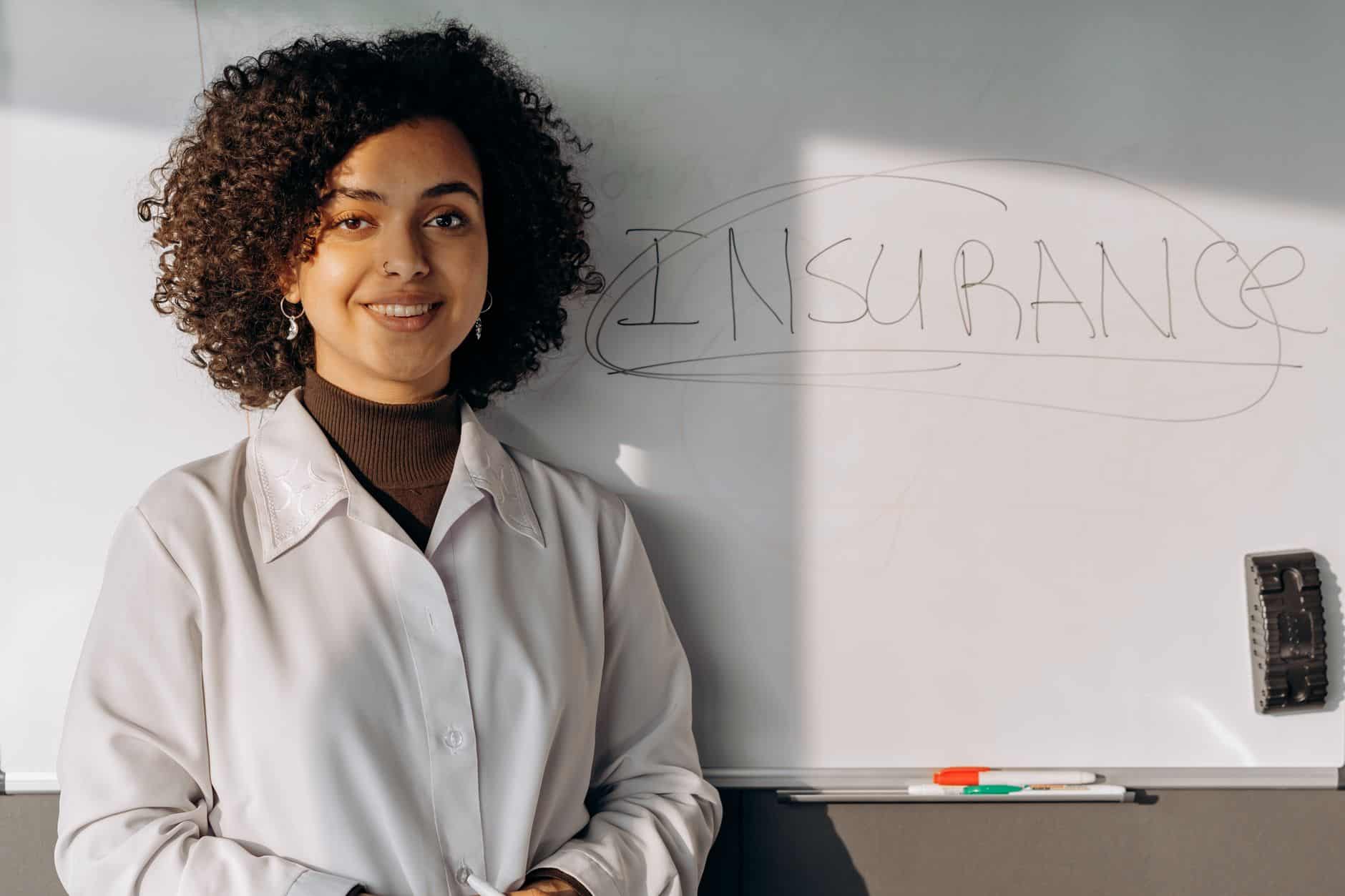 African American woman smiling in office setting with a whiteboard displaying 'Insurance'.