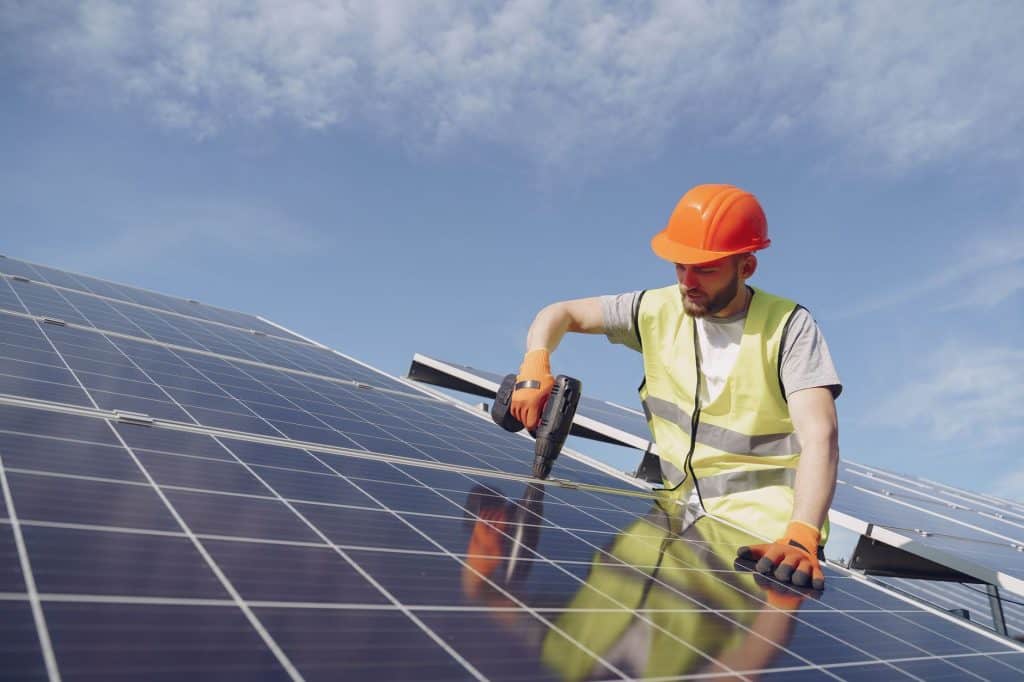 From below of concentrated young bearded male technician in protective clothes mounting solar batteries using drill agai