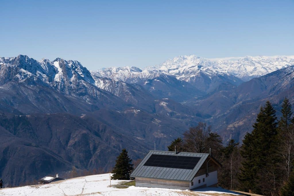 A stunning alpine view of snowy mountains and a solar-powered cabin under a clear blue sky.