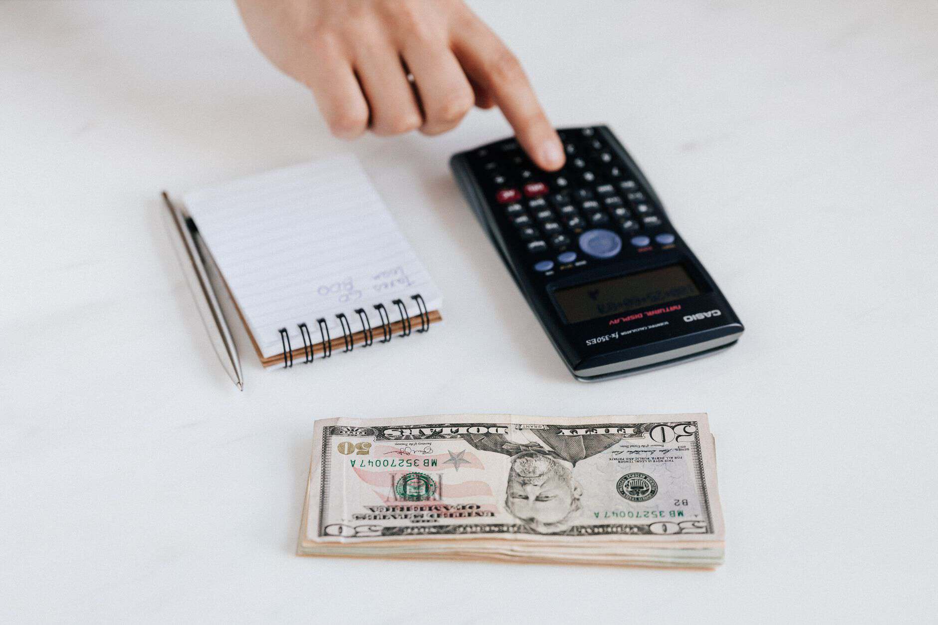 Top view of crop faceless economist touching calculator button while counting income near heap of paper bills and small