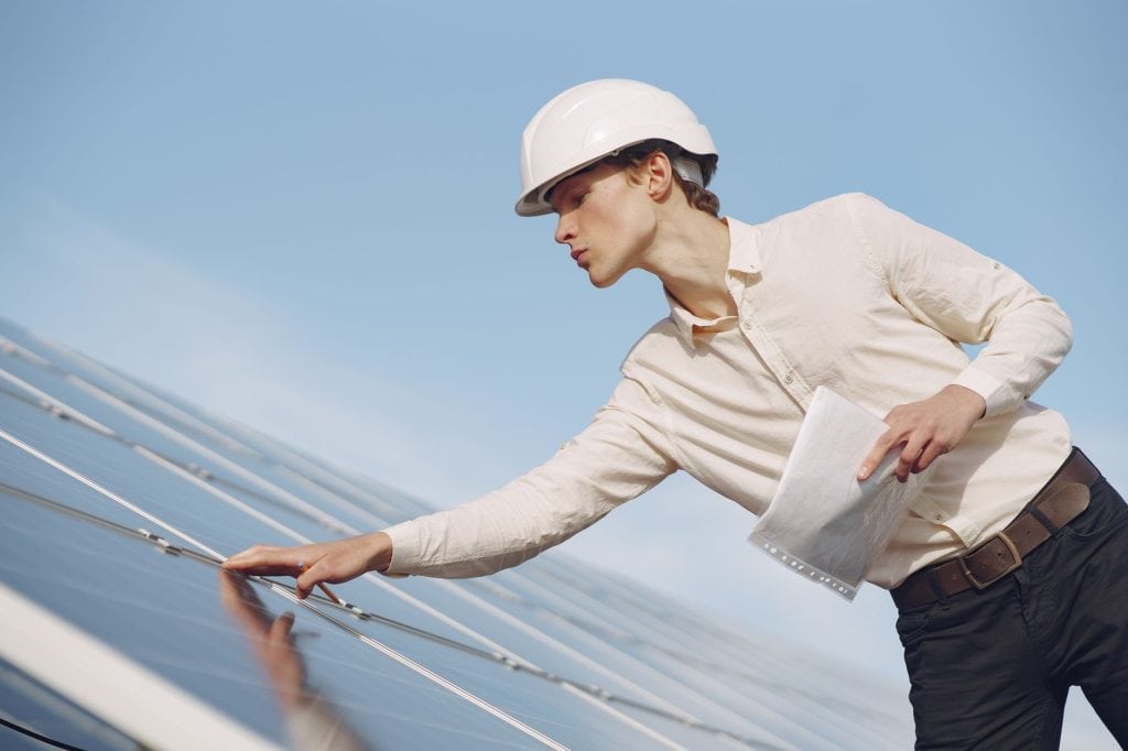 Low angle of young male inspector with documents in formal wear and hardhat standing against solar panels and checking d